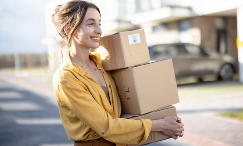 A woman carrying three stacked cardboard boxes on a clear day.